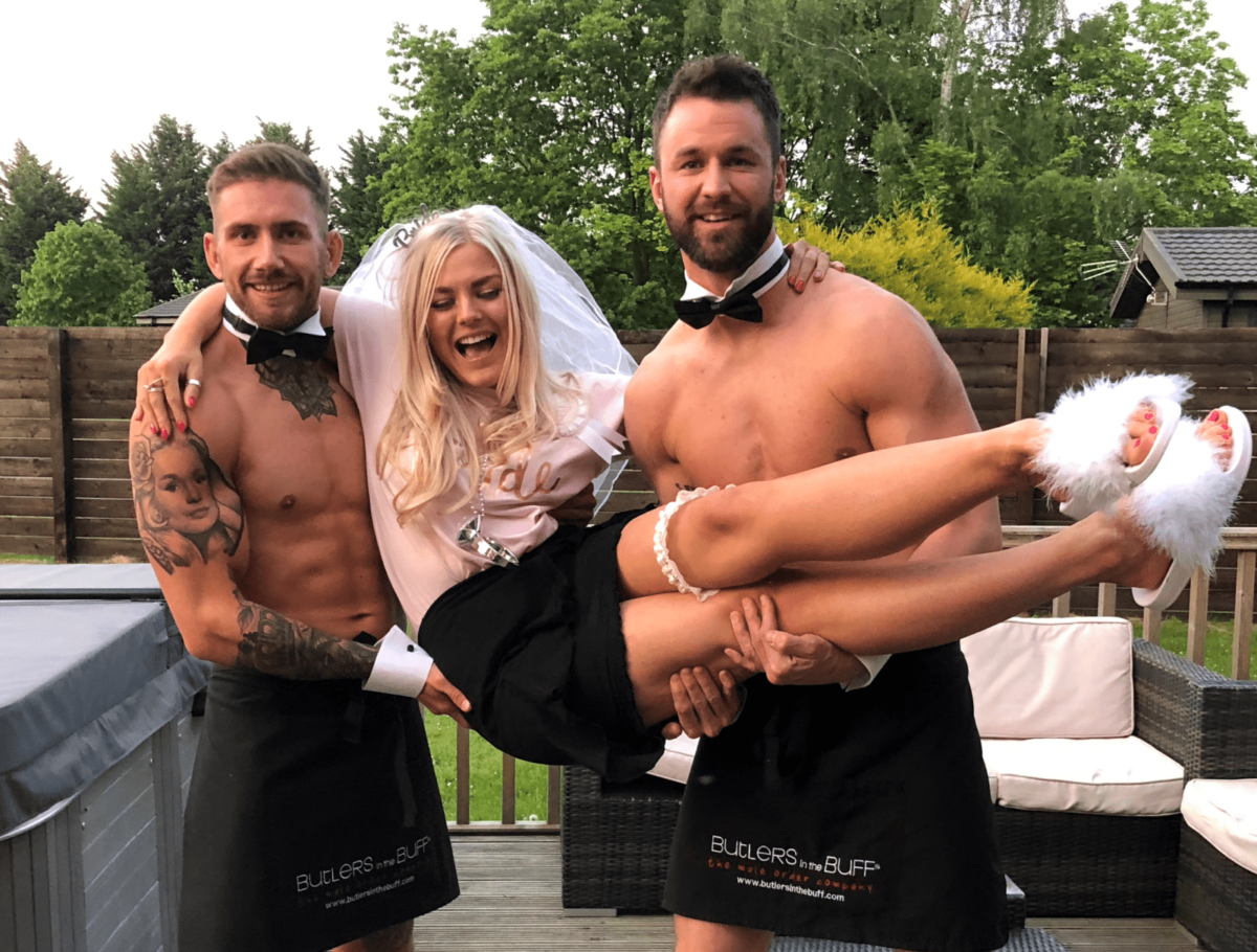 Bride-to-be smiling and being lifted by two Buff Butlers wearing aprons and bow ties during a fun hen party on a garden patio.