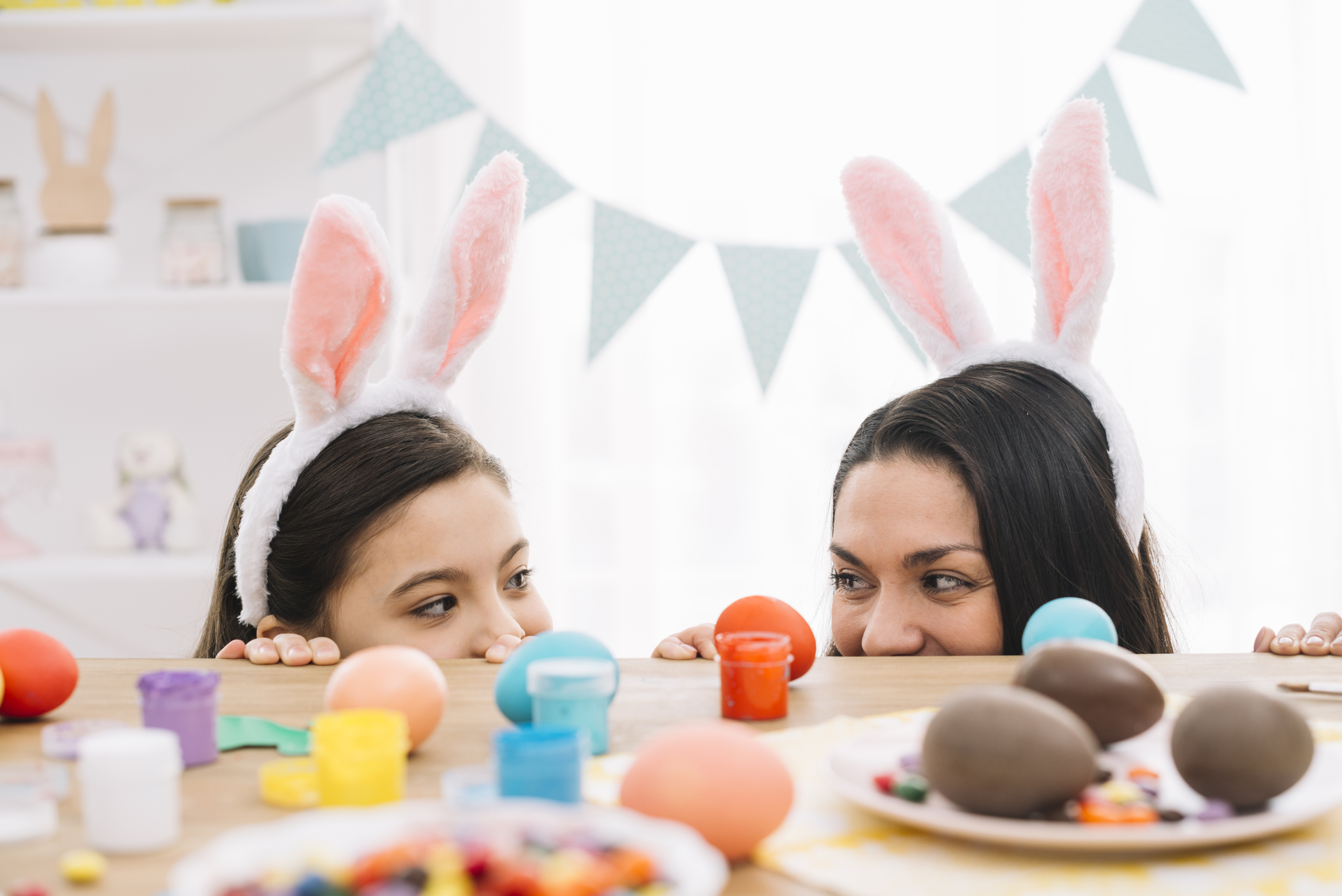 UTF-8''mother-daughter-with-bunny-ears-peeking-out-from-table-with-tasty-easter-eggs.jpg